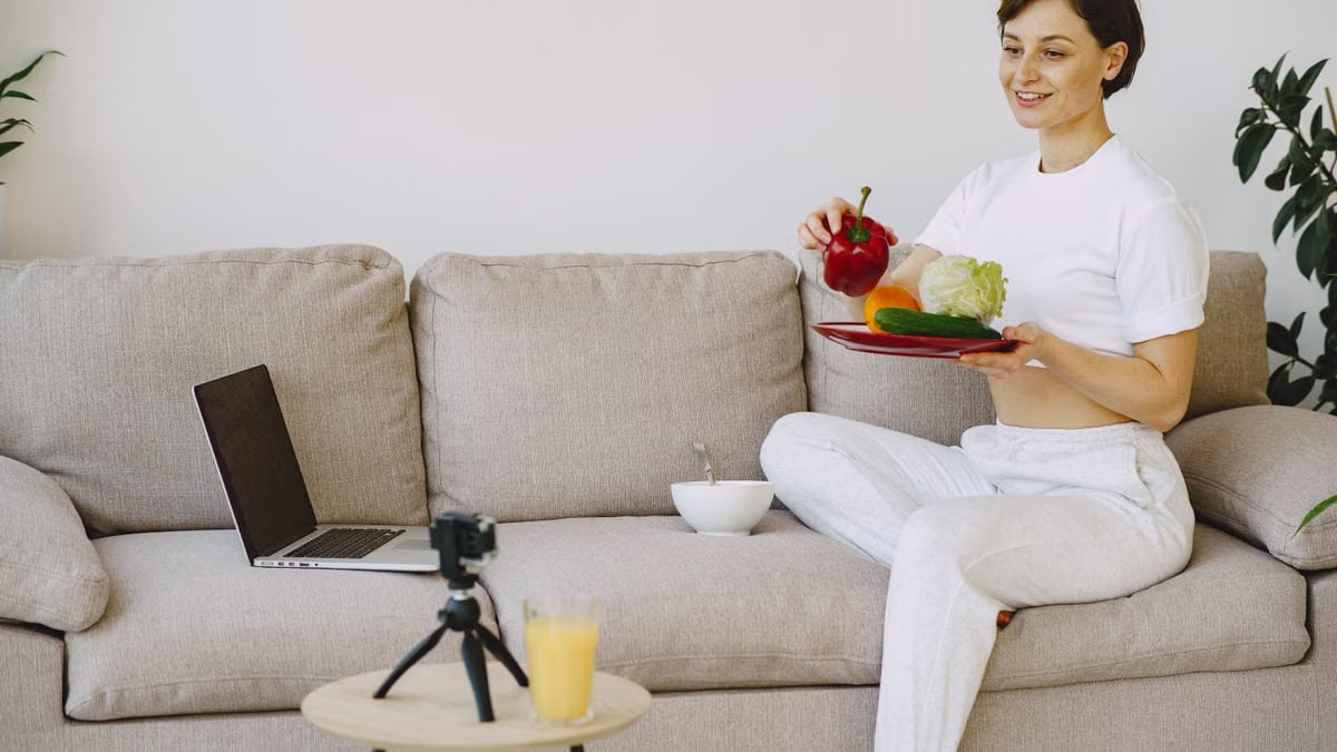 Content female in white outfit sitting on sofa holding plate with healthy food in hand and taking photo on camera